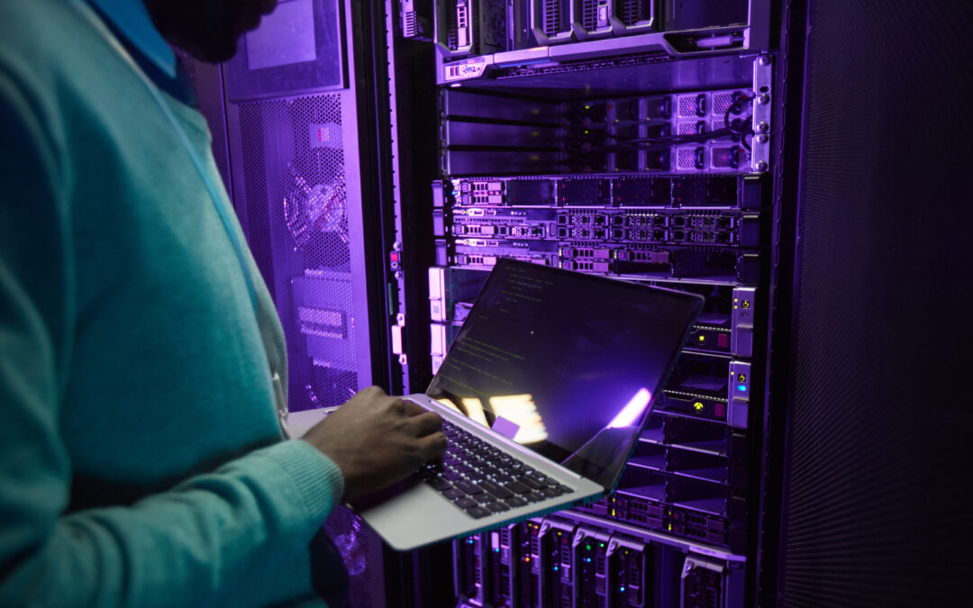 A person stands in a server room while using a laptop.